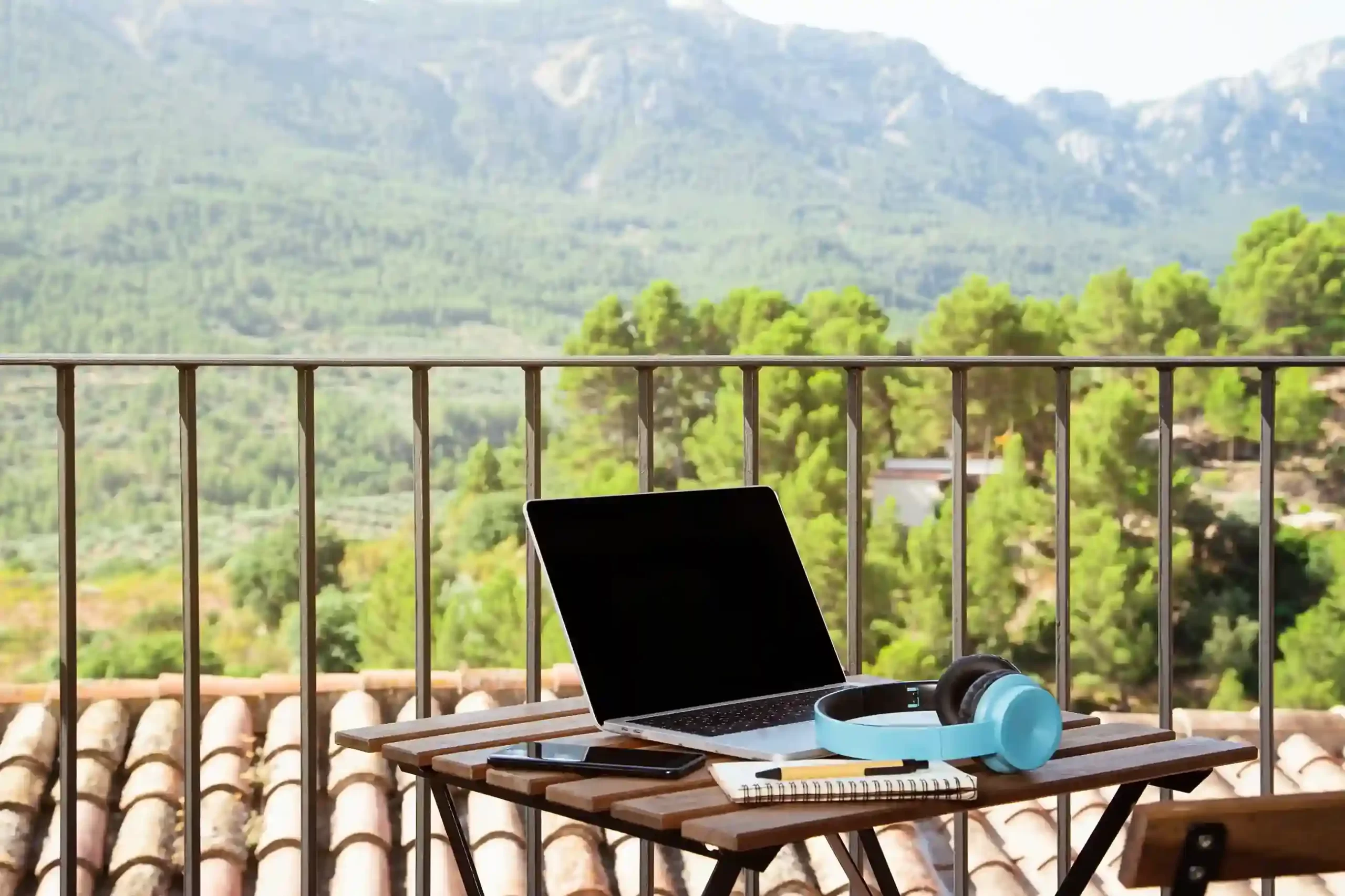 Laptop, blue headsets, a notebook, and a cellphone on the table on a balcony with a beautiful view
